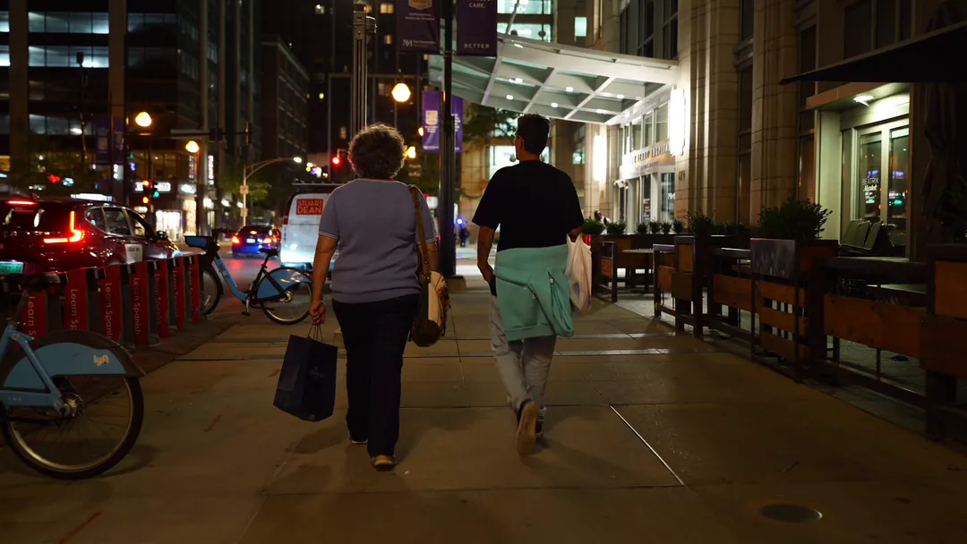 Two people walk a downtown sidewalk at night past lit restaurant patios.