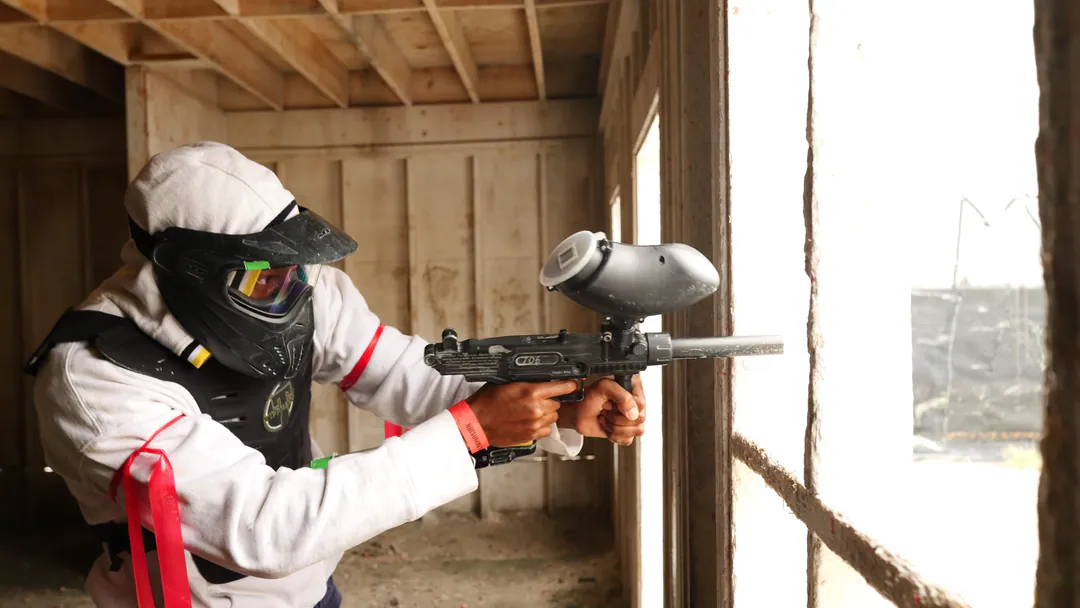 A paintball player in a white mask aims from the doorway of a wooden barn.
