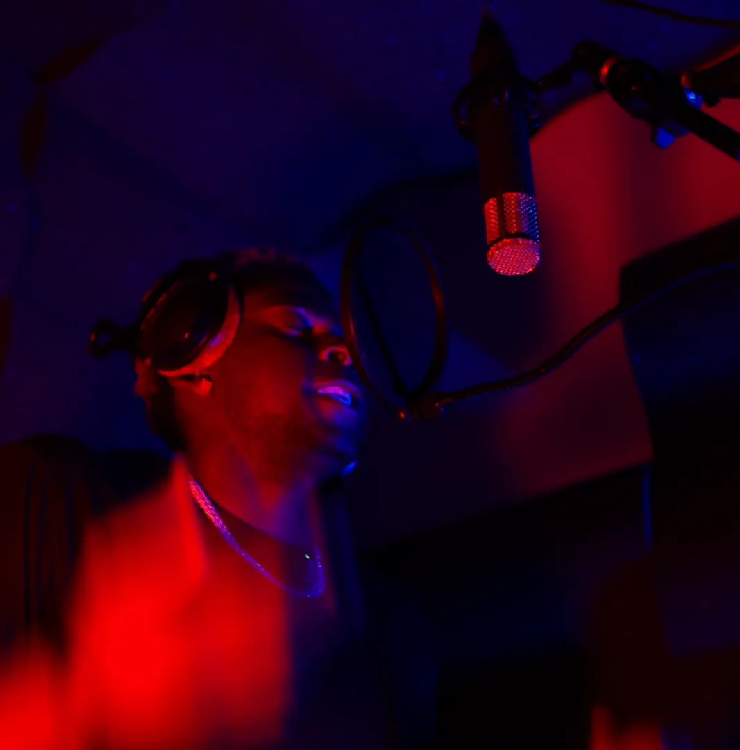 An artist records in a vocal booth under red and blue light, headphones on, mic overhead.