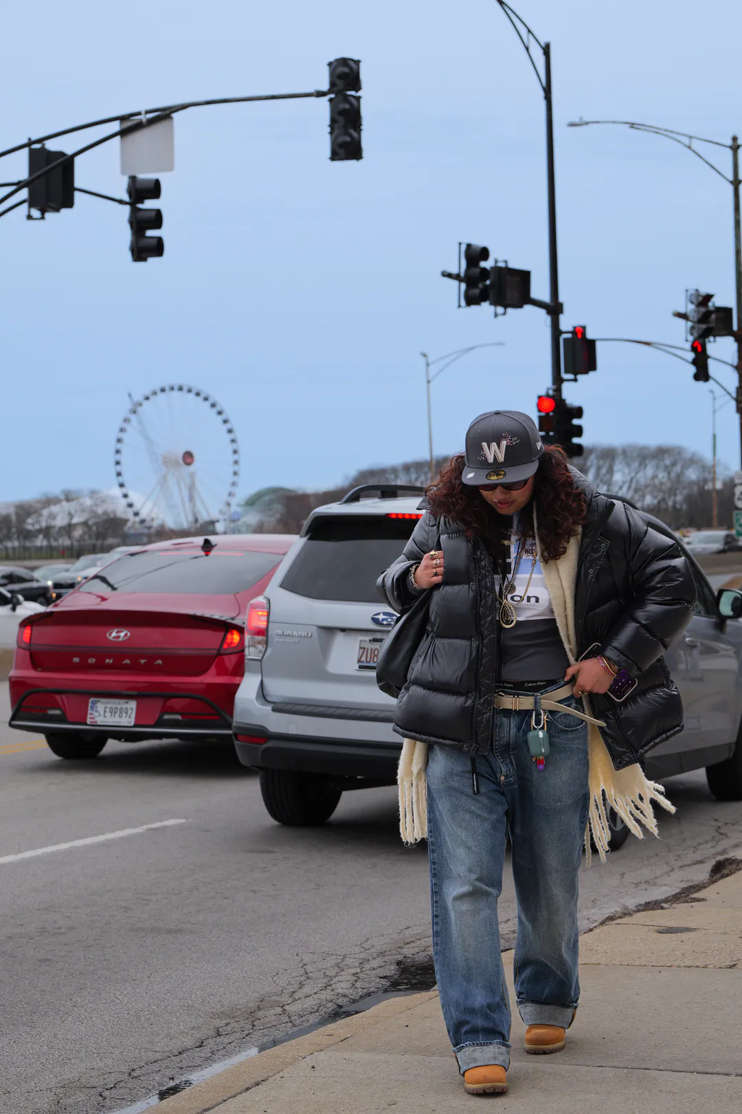A woman in a black puffer jacket and wide-leg jeans walks along a Chicago street, the Navy Pier wheel behind her.