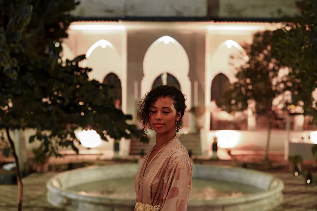 A woman in an embroidered kaftan stands in a lit courtyard at night, arched doorways and a fountain behind her.