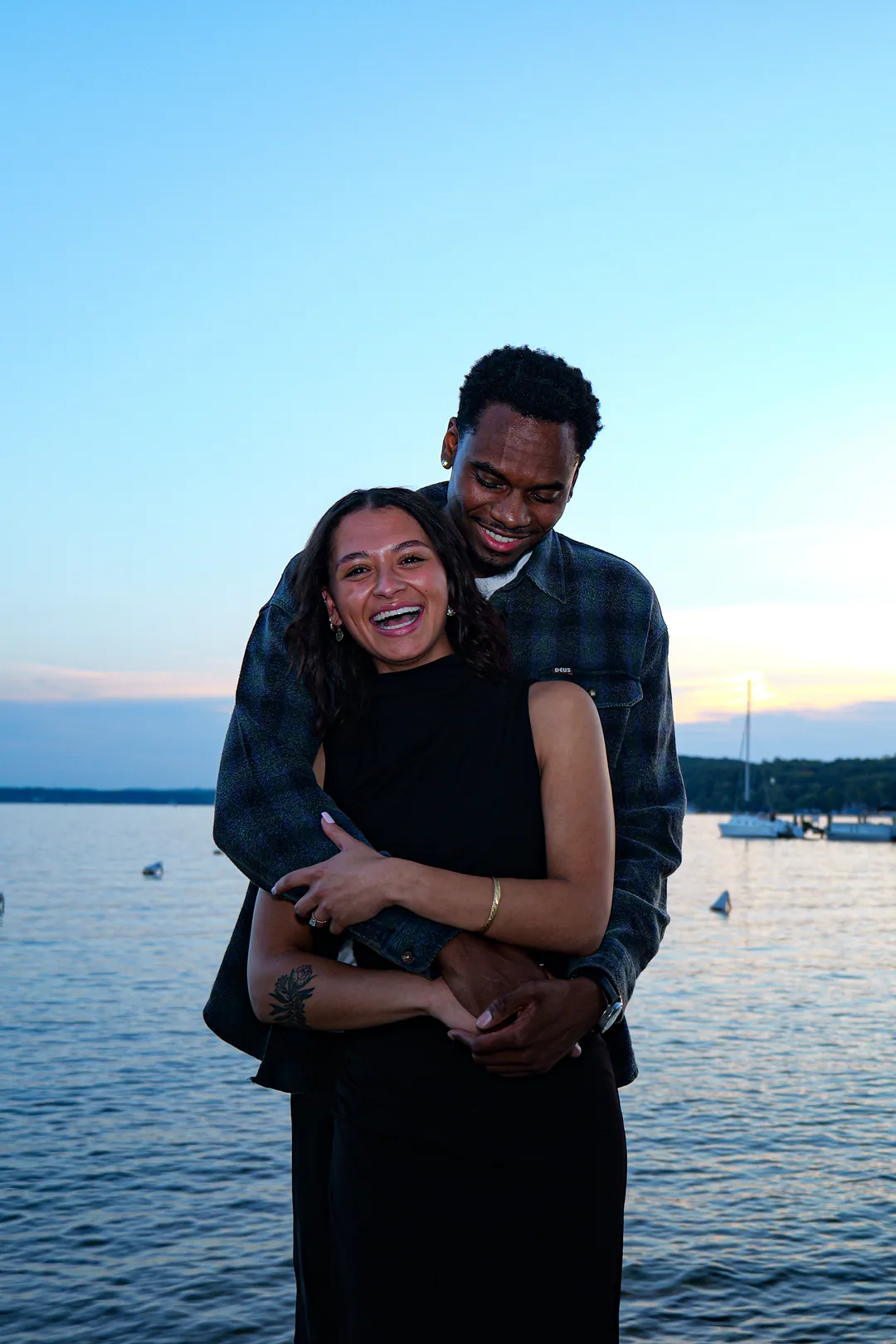 A couple embraces by the water at dusk, a sailboat moored behind them.