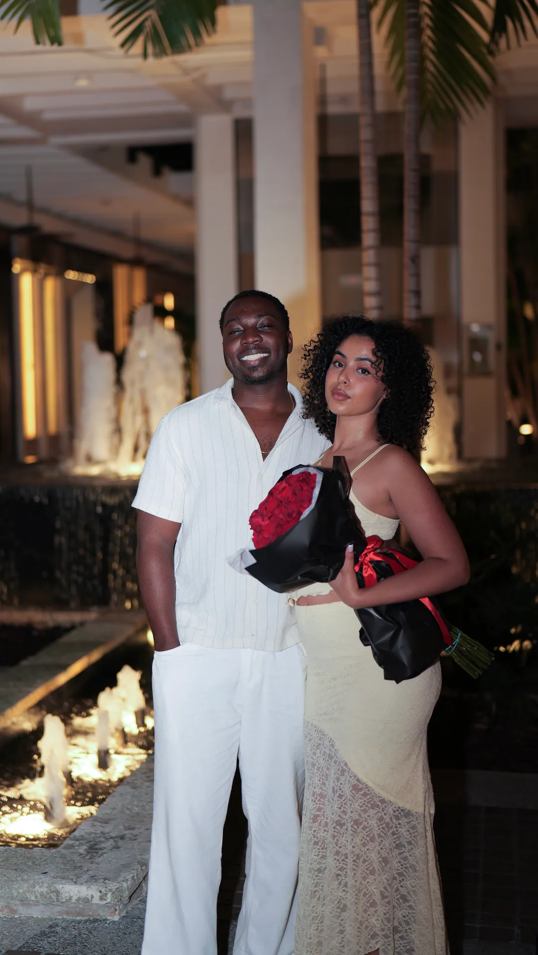 The newly engaged couple at night beside a lit fountain, holding a bouquet of red roses.